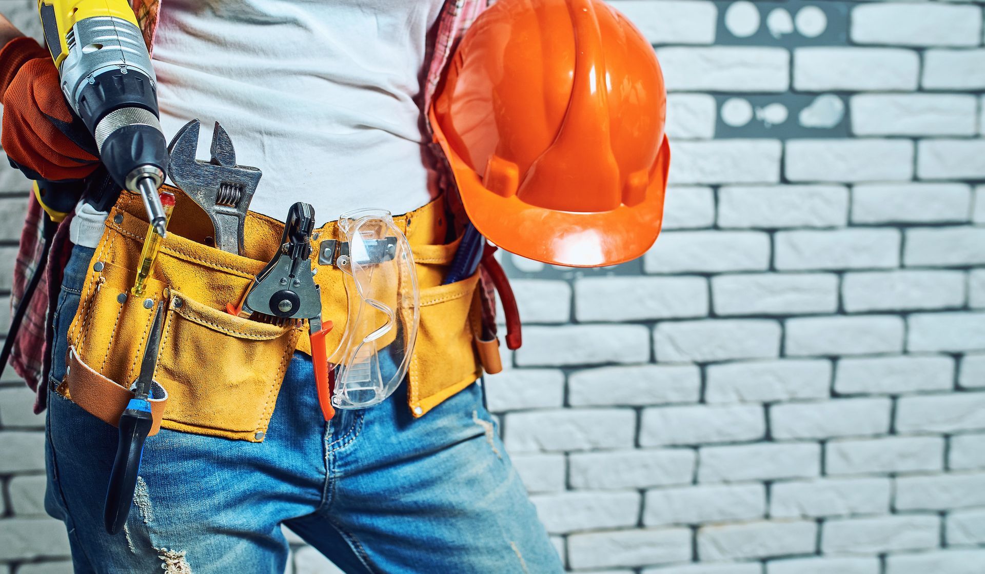 Operaio edile con cintura portautensili, casco e trapano in mano, di fronte a un muro di mattoni.