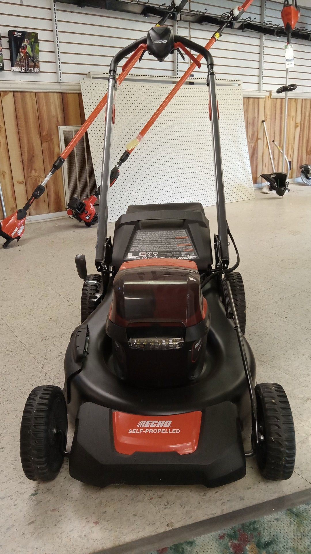 A black and orange EGO Power+ cordless lawn mower sits on a grey floor in a retail store with tools in the background.