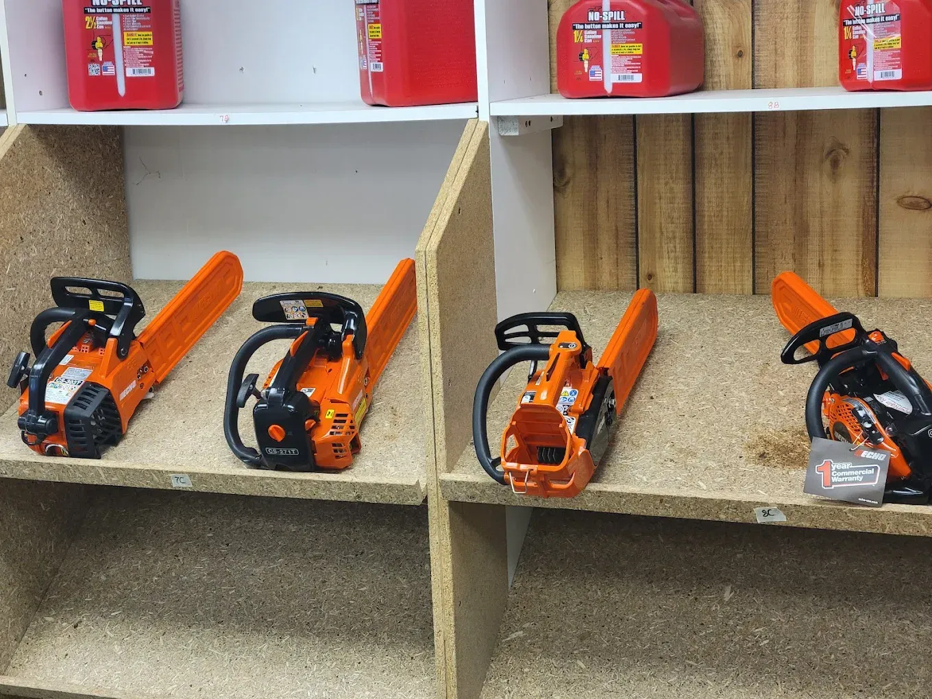 Four orange chainsaws arranged on shelves in a retail display, with red fuel cans stored on the shelf above.