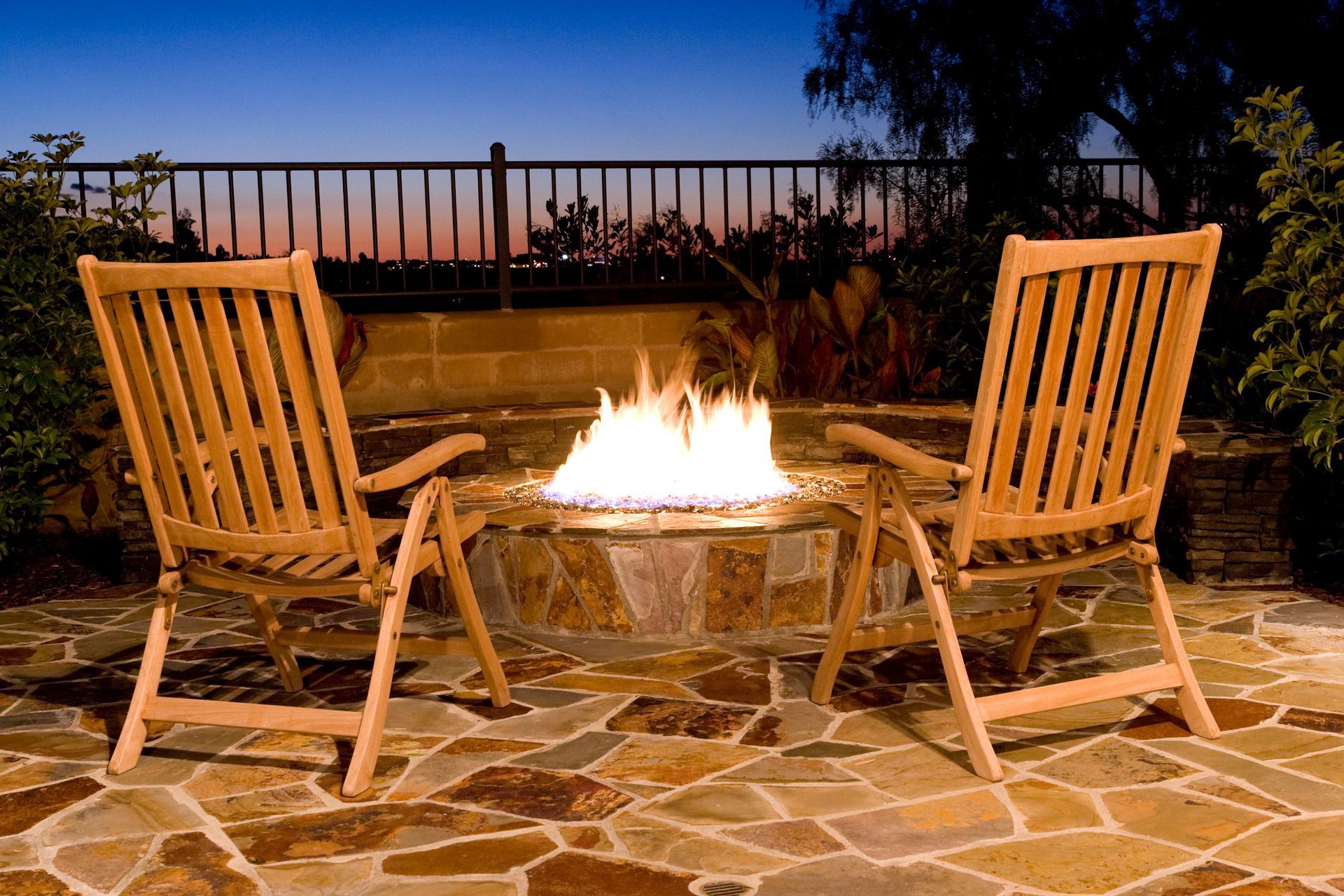 Two wooden chairs face a fire pit on a stone patio at dusk.