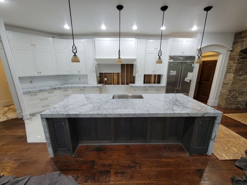 Kitchen under construction: white cabinets, gray island with marble countertop, wood floor, and hanging lights.
