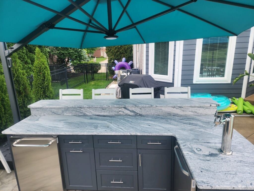 Outdoor kitchen with blue granite countertop, cabinets, and tap; grill under a teal umbrella.