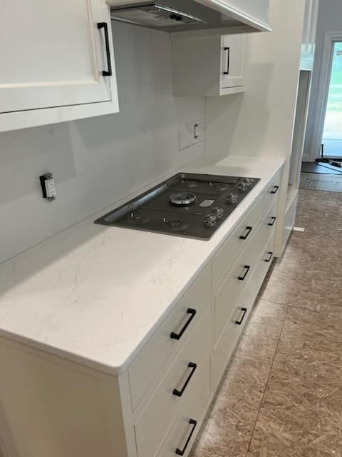 White kitchen cabinets and countertop with a built-in stove; black hardware; unfinished floor.