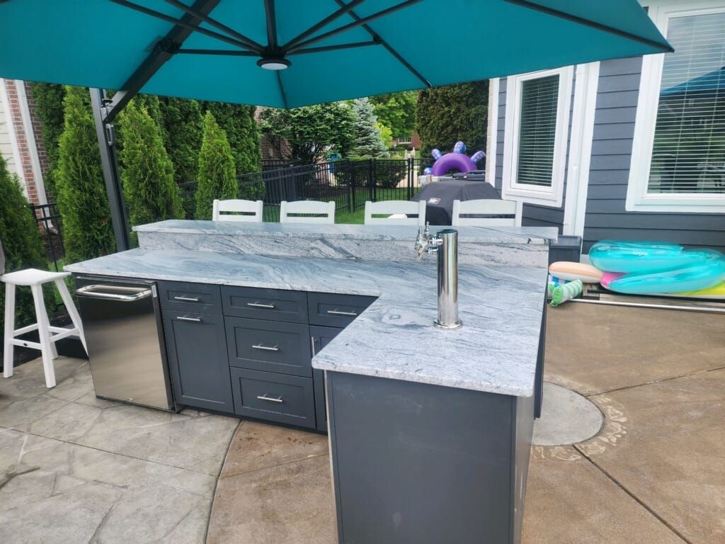 Outdoor kitchen with gray cabinets, granite countertop, beer tap, under blue umbrella.