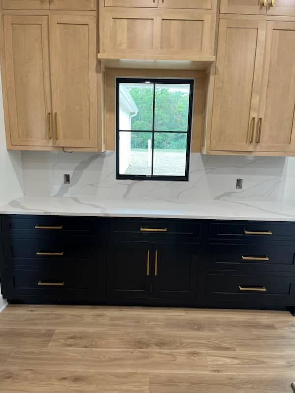 Black and tan kitchen cabinets with gold hardware, white marble countertops, and a window.