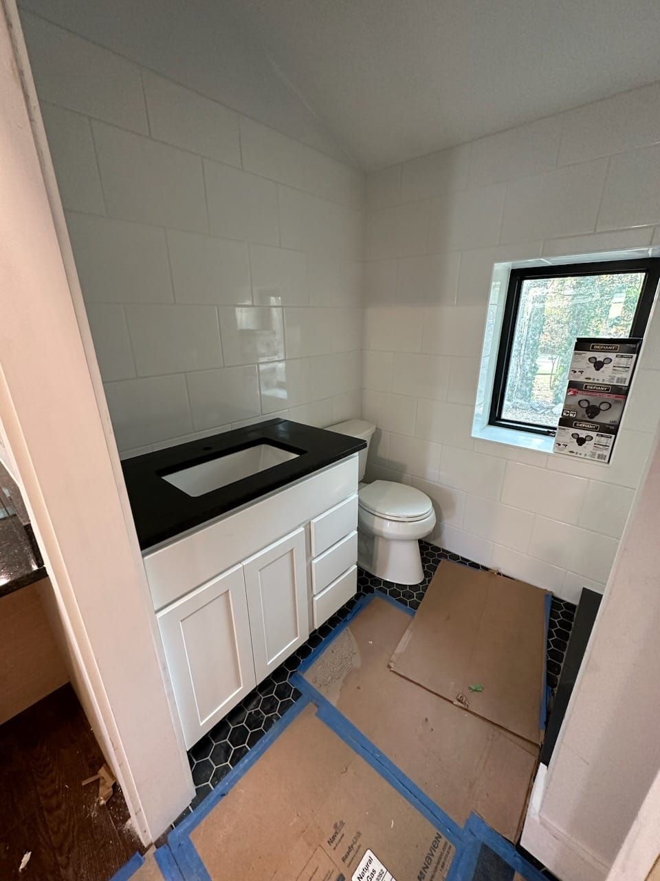Small bathroom with white tiled walls, white vanity with black countertop, toilet, and small window.