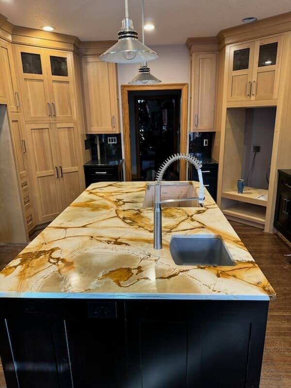 Kitchen island with golden-veined countertop, stainless steel faucet, dark base. Light wood cabinets surround. Kitchen island with golden-veined countertop, stainless steel faucet, dark base. Light wood cabinets surround.