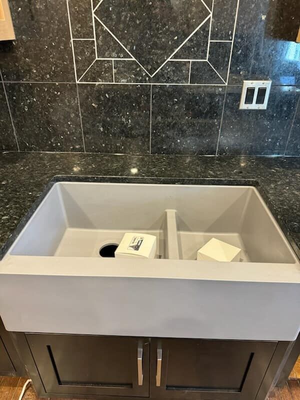Gray double-basin sink in a dark-countertop kitchen. Dark cabinets below. Black speckled backsplash with geometric accents.