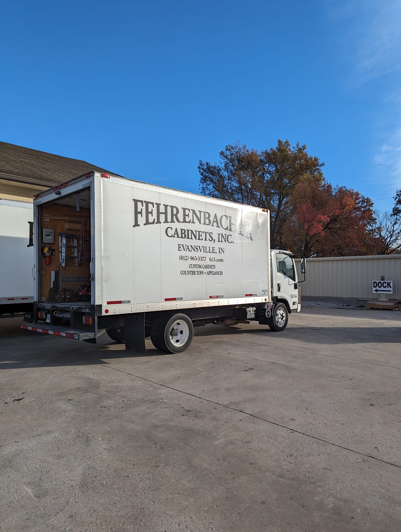 White moving truck with open back, parked in gravel lot on a sunny day.
