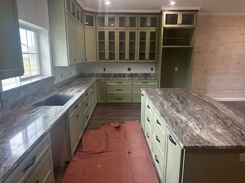 Green kitchen cabinets with granite countertops, island, and sink by a window.