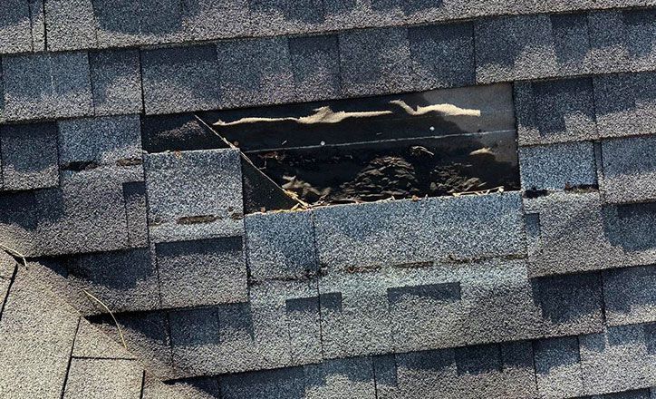 Damaged asphalt roof shingles with a large hole revealing the underlayment.