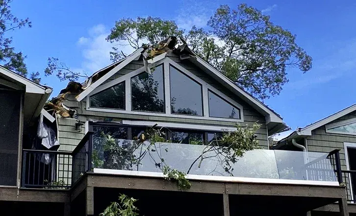 House with roof damage, broken windows, and debris on balcony after a storm; blue sky.