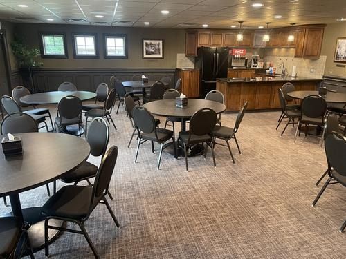 Empty community room with round tables and black chairs, buffet counter, and wood-paneled wall with windows.