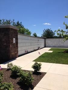 Sunny backyard patio with concrete walkway, stone pillar, white fence, and small shrubs along a garden bed.