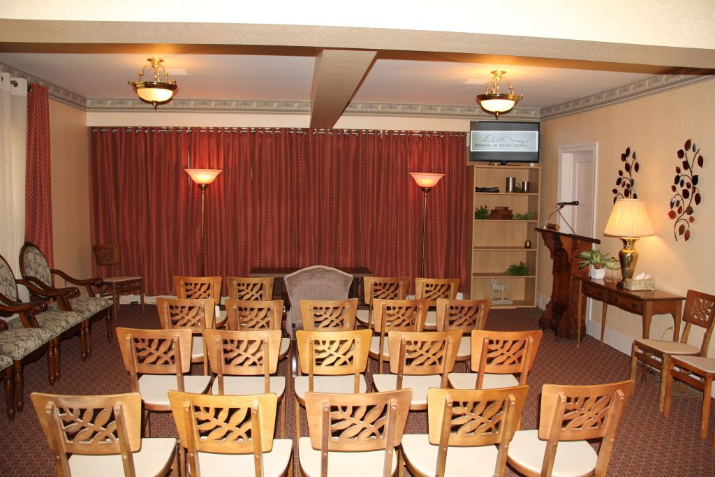 Interior of a room arranged for a service, with rows of chairs facing a red curtain. Walls are cream-colored, and decorative sconces and light fixtures are present.