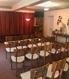 A funeral home chapel with chairs facing a raised platform with red curtains. There's a sideboard, lamps, and a bookshelf in the background.