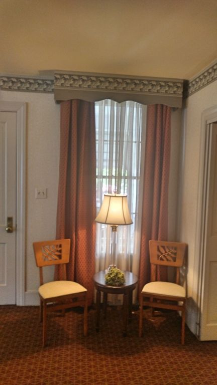 Two chairs face a small table with a lamp in front of a window with pink curtains. The scene is in a room with dark red carpet.