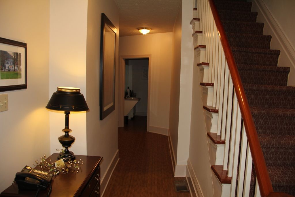 A hallway with a staircase on the right and a small table with a lamp and telephone. A door is visible at the end of the hallway.