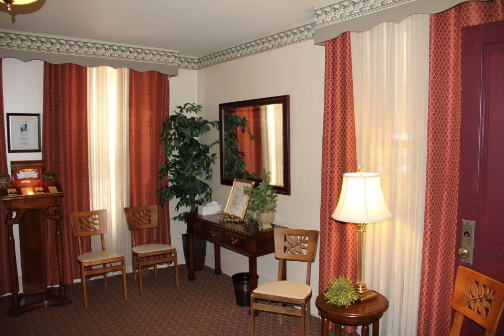 A warmly lit waiting area with red and cream curtains, chairs, and a table. A mirror and plants add to the decor.