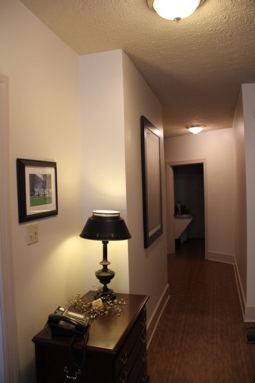 Narrow hallway with wood floor, white walls, and a dark wooden table with a lamp and a framed picture.