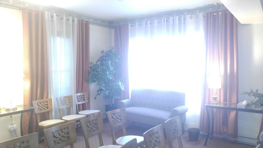 A bright room with rows of chairs, a sofa, and large windows with curtains. A large potted plant sits in the center.