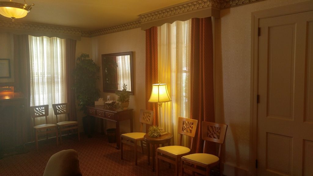 A dimly lit room with three chairs, a table, and a mirror, likely a waiting room. Orange curtains and warm lighting create a subdued atmosphere.