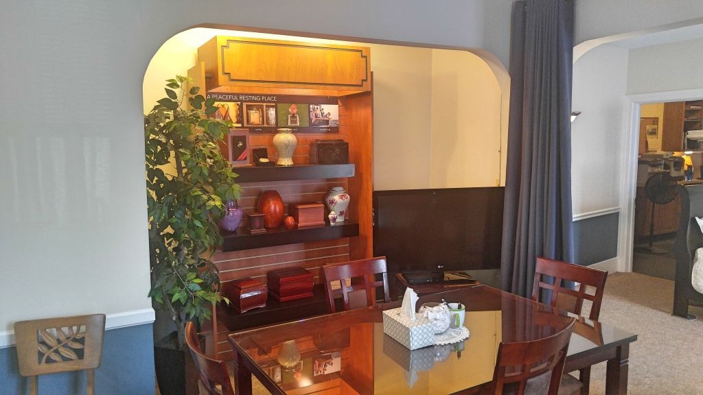 A dining room with a wooden table and chairs, a built-in bookshelf displaying decor, and a large artificial plant.