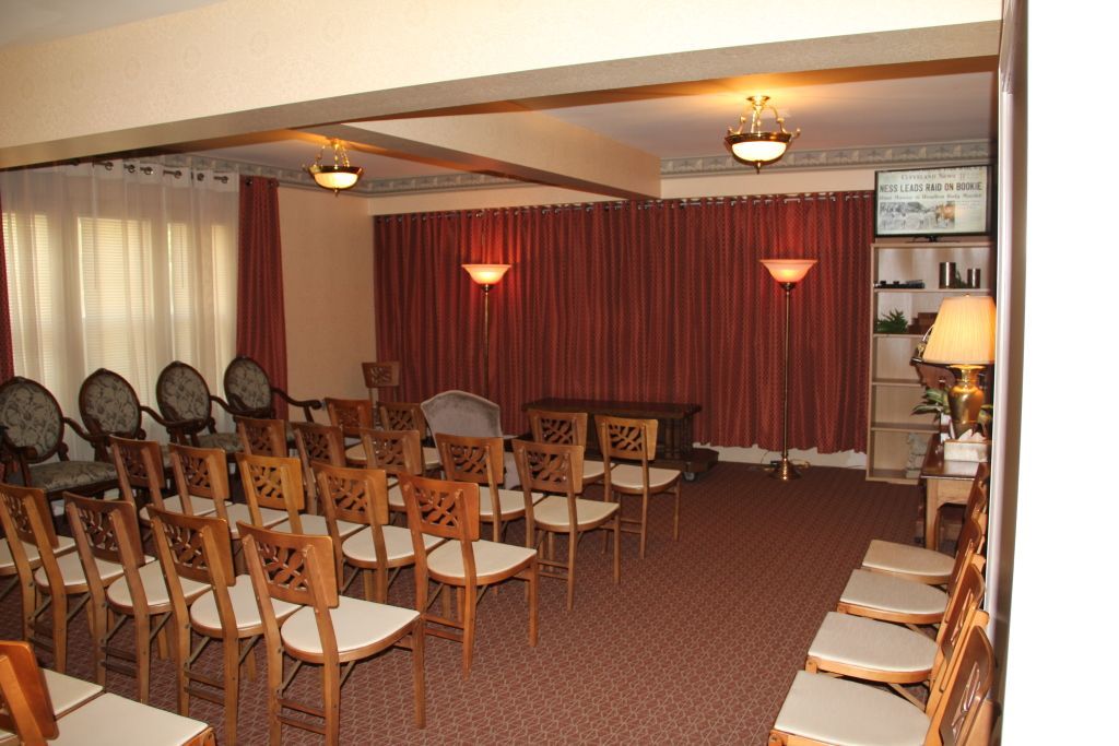 A funeral home chapel with rows of wooden chairs facing a closed casket and red curtains; soft lighting and beige walls.
