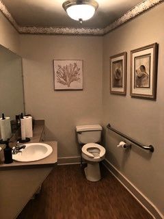 A small bathroom with a toilet, sink, and grab bar. Artwork and a paper towel dispenser are visible. The walls are beige, and the floor is wood-like.