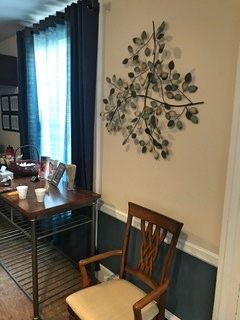 A room with a wooden chair, table, and a metal leaf wall art piece on a tan wall next to a window with blue curtains.