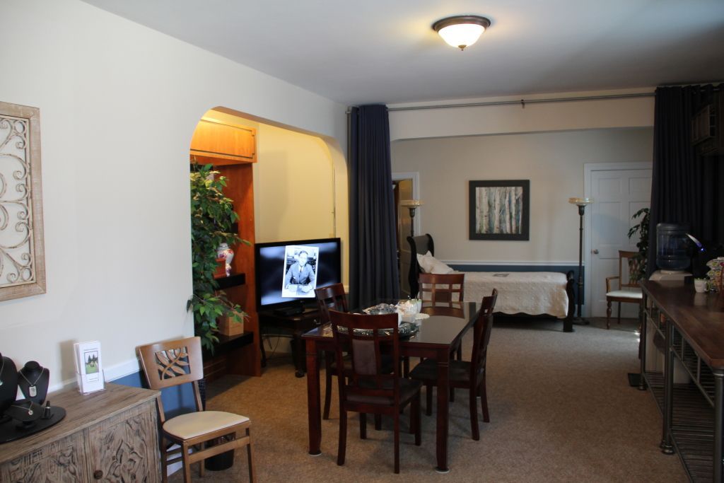 An open-concept living space with a dining area, TV, and bed visible behind a curtain. Beige walls, dark wood furniture, and a few decorative plants.