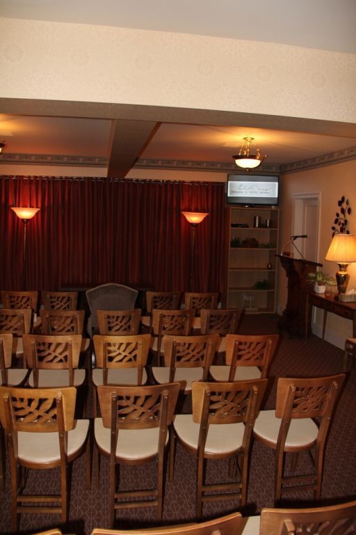 Rows of wooden chairs face a raised platform with a lectern and screen. A red curtain covers the back wall in a dim room.