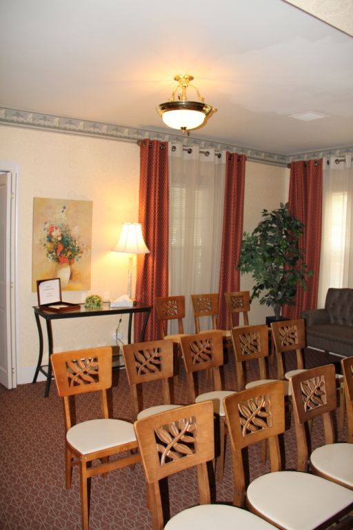 A small room set up for a gathering, with wooden chairs arranged in rows, a table with flowers and a painting, and burgundy curtains.