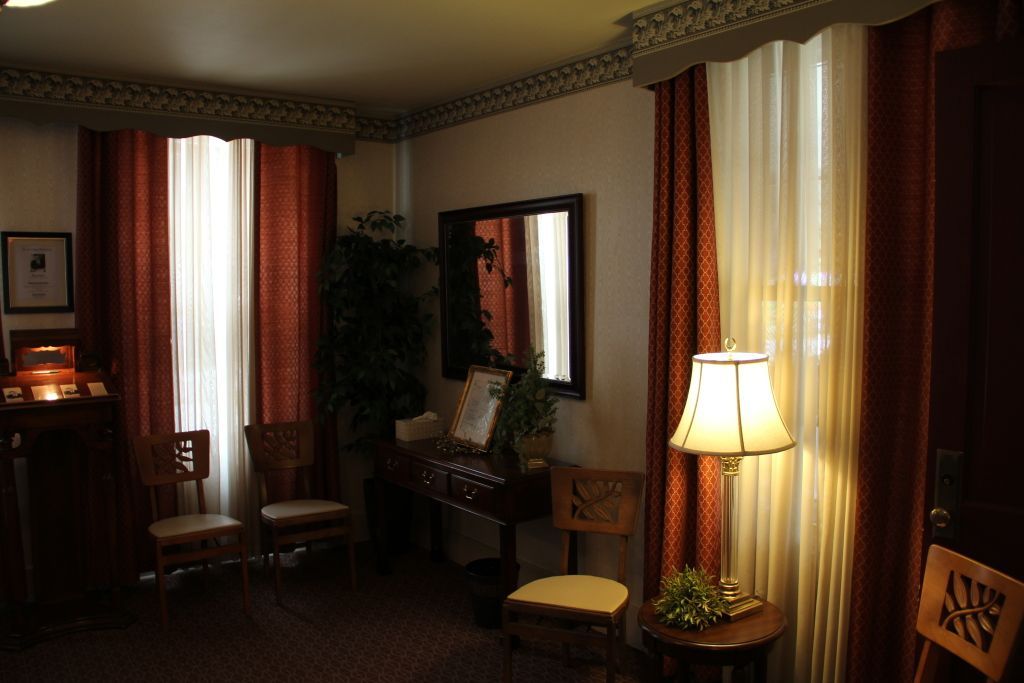 An interior room with two windows with red and white curtains, a mirror, and a lamp. Chairs and a table are in the room.