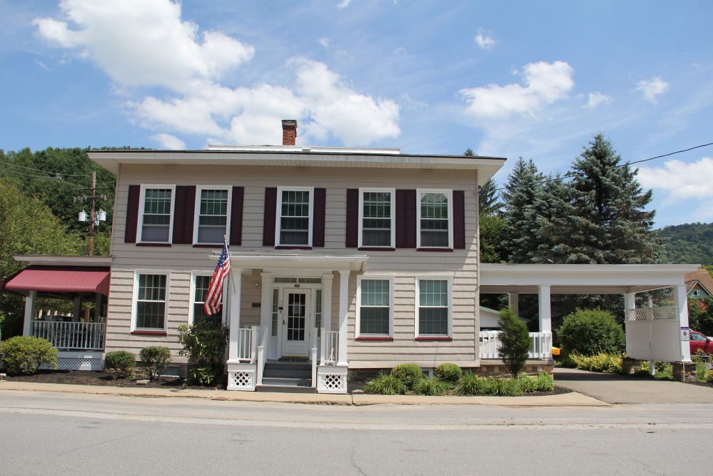 Two-story, beige house with maroon shutters, a porch, and a carport, an American flag, and a red awning under a blue sky.