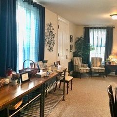 A living room with blue curtains, a long wooden table with various items, two armchairs, and a houseplant.