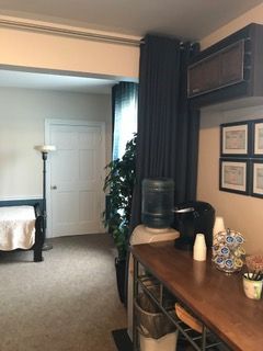 A small office break area with a water cooler, coffee machine, and microwave. A doorway leads to a room with a bed and lamp.