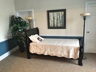 A bedroom featuring a dark-framed bed with a light-colored quilt. A painting hangs above, and two floor lamps flank the bed.