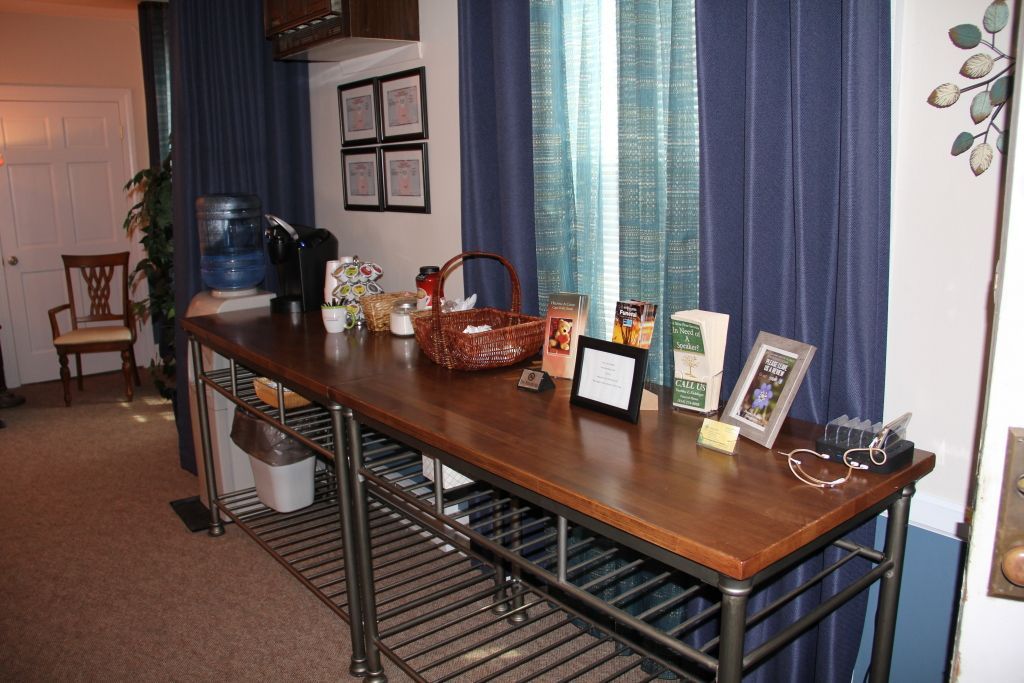 Long wooden table with items on top, in front of blue curtains. A water cooler, basket, and framed pictures are visible.