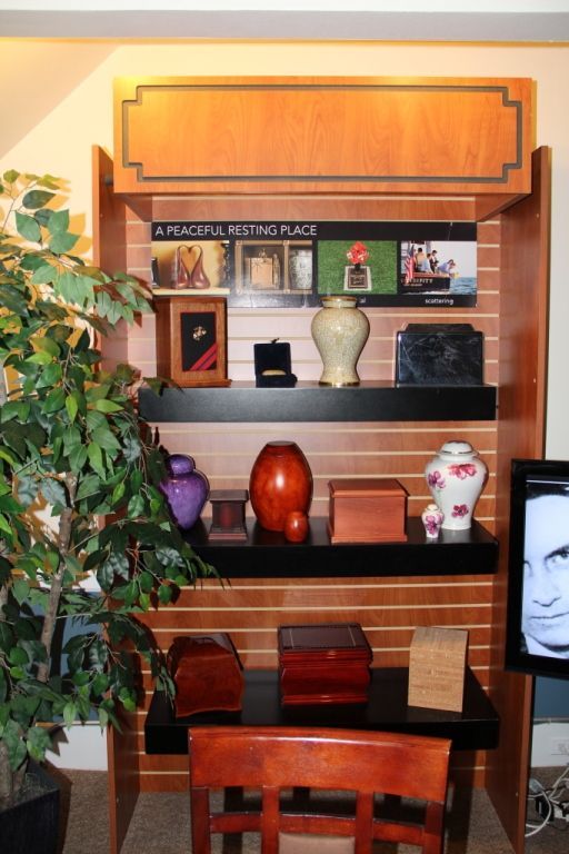 Display of memorial urns and containers on shelves, with a plant and a person's portrait nearby. The sign reads 