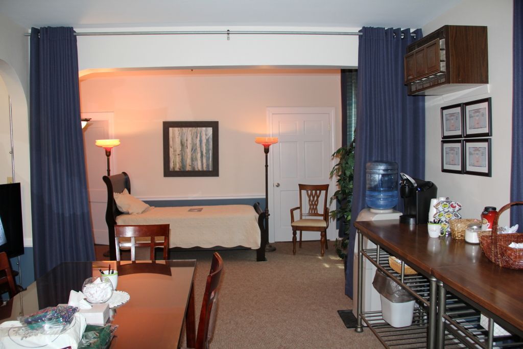 An open-concept living space with a bedroom area in the background, featuring a bed, artwork, and floor lamps. A dining table, kitchen counter, and blue curtains divide the space.