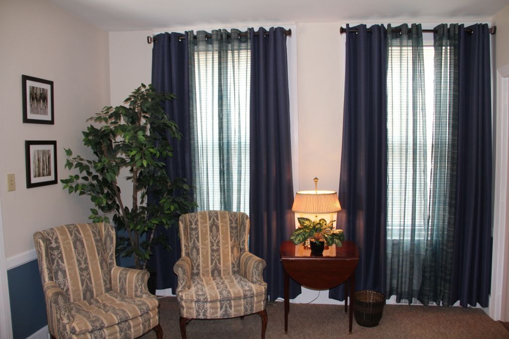 Two armchairs and a small table sit in front of two windows with blue curtains. A large plant is next to the chairs.