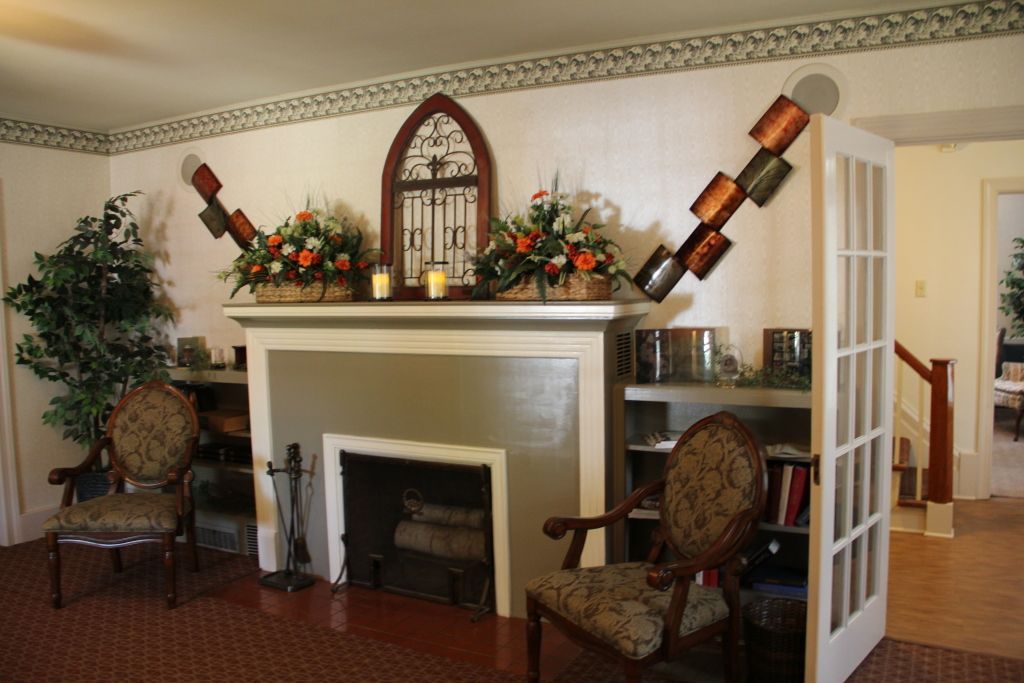 A fireplace is flanked by two chairs, bookshelves, and decorative metal shovel sculptures. Above the mantle are floral arrangements and a decorative arched wooden piece.