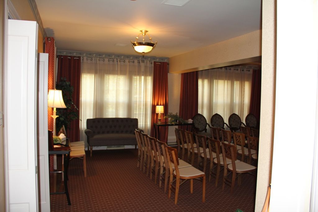 Interior of a funeral home chapel with rows of chairs facing a sofa and windows with red curtains.