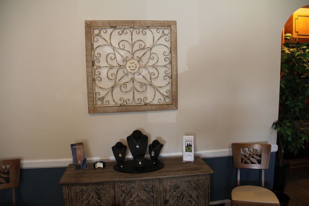 A wall with decorative metal art, a wooden cabinet displaying jewelry, and a chair. A second chair is visible on the left.