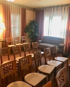 A room with wooden chairs arranged for a gathering, pink drapes, a couch, and a large plant by a window.