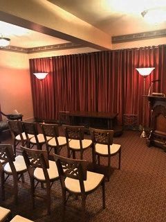 A room set up for a service with rows of chairs facing a dark wooden table, red curtains, and wall sconces.