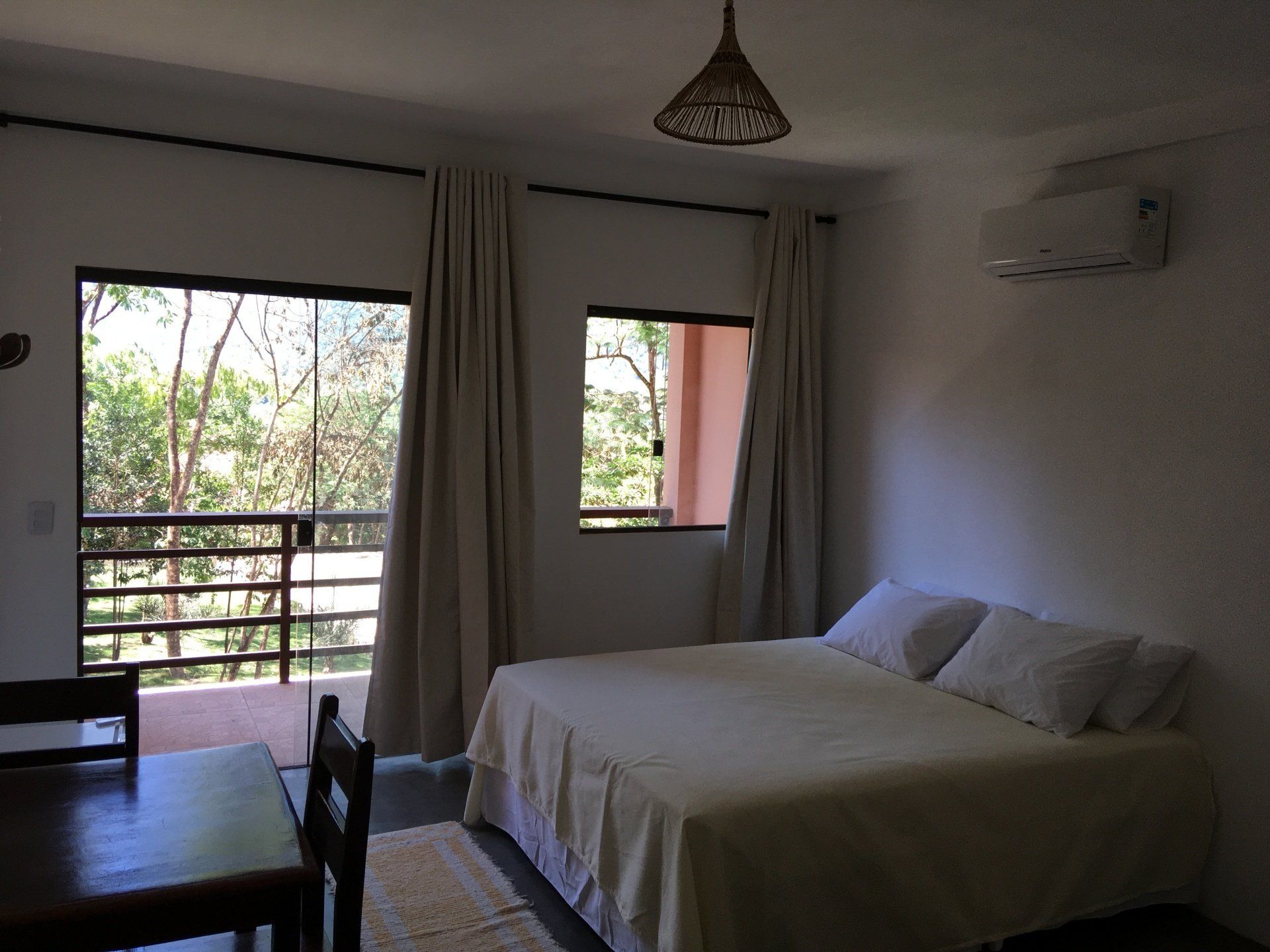 Quarto com cama, janelas, mesa e cadeira, cortinas e ar condicionado.