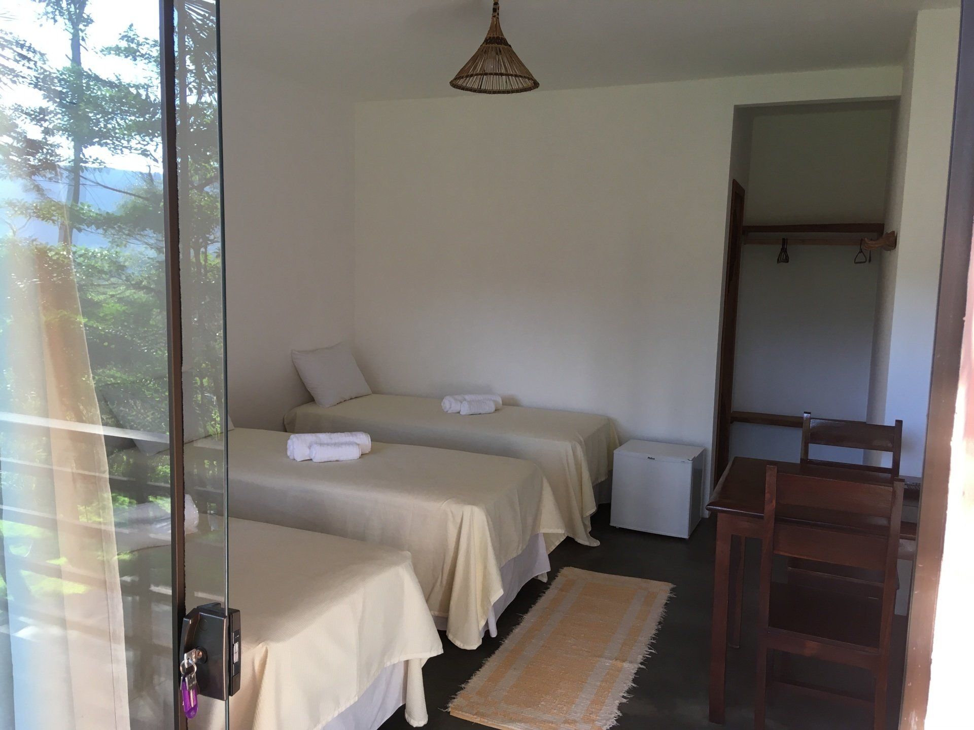 Quarto com três camas de solteiro, geladeira pequena e escrivaninha de madeira, com vista para as árvores através de uma porta de vidro.