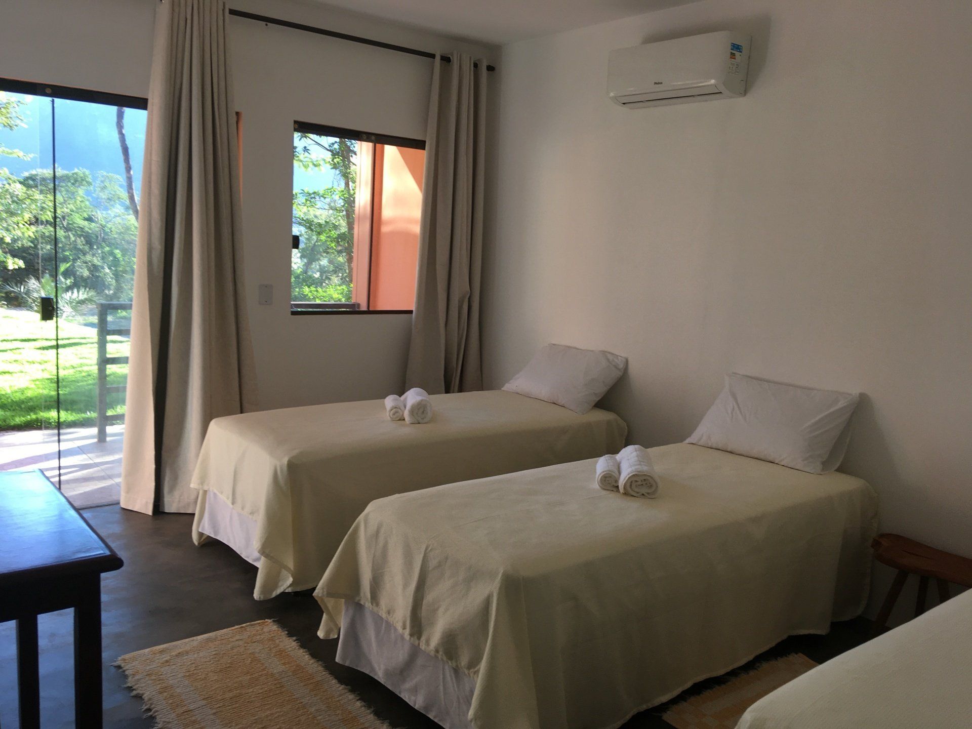 Três camas de solteiro em um quarto iluminado com janela e porta de vidro, lençóis creme e paredes brancas.